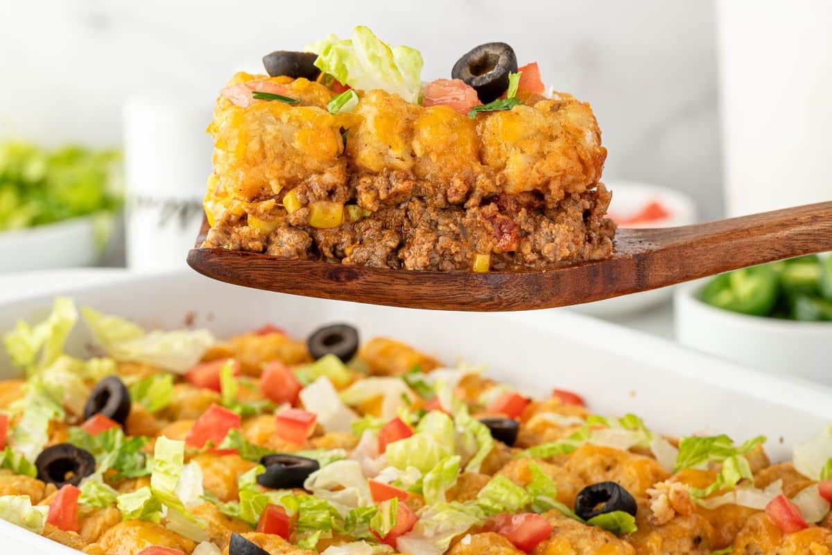 A wooden spoon holds a serving of taco casserole topped with cheese, lettuce, tomatoes, and black olives.