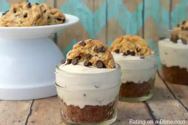 Mini mason jars with cookie crust, creamy filling, chocolate chips, and a cookie on top; cookies on a stand behind.