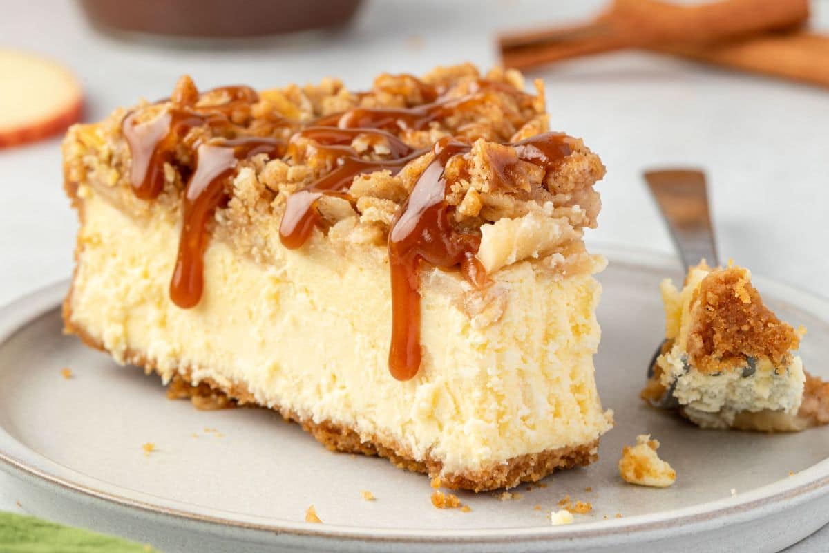 A slice of cheesecake topped with crumbly streusel and caramel sauce on a plate with a forkful beside it.