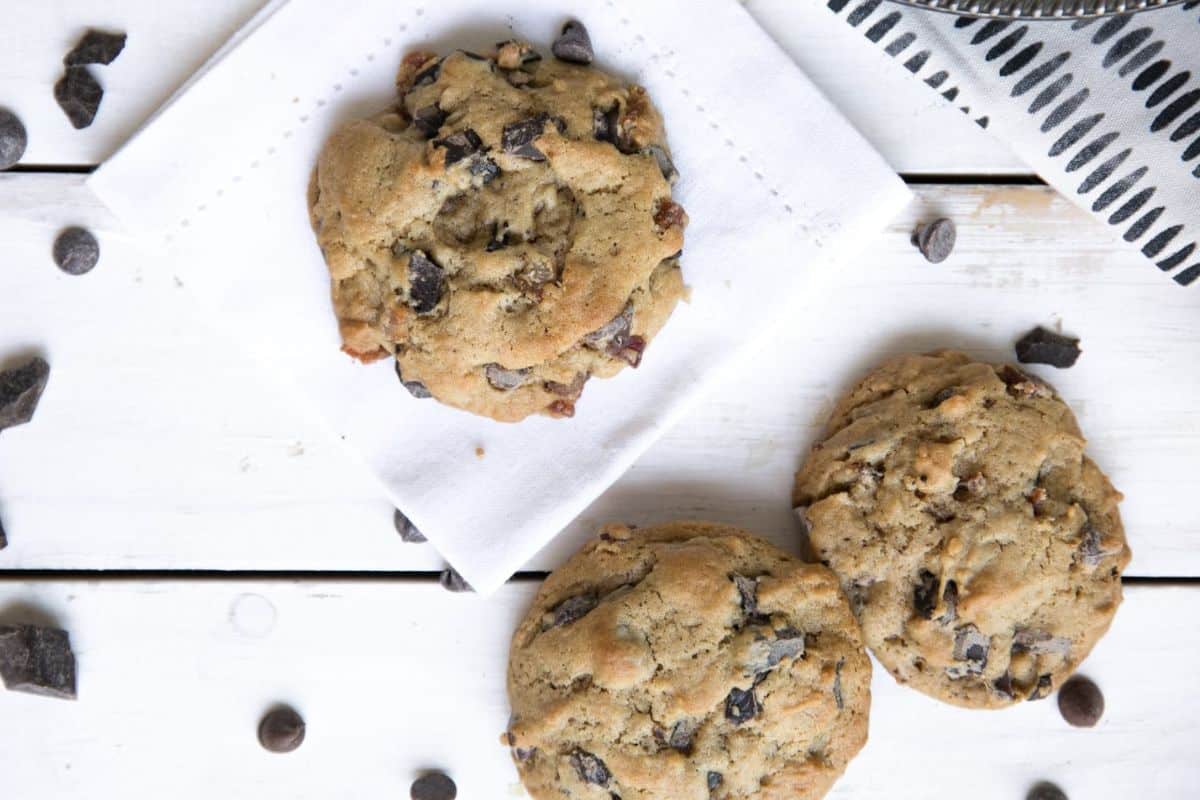 Three chocolate chip cookies on a white wooden surface, one on a folded napkin, with scattered chocolate chips nearby.