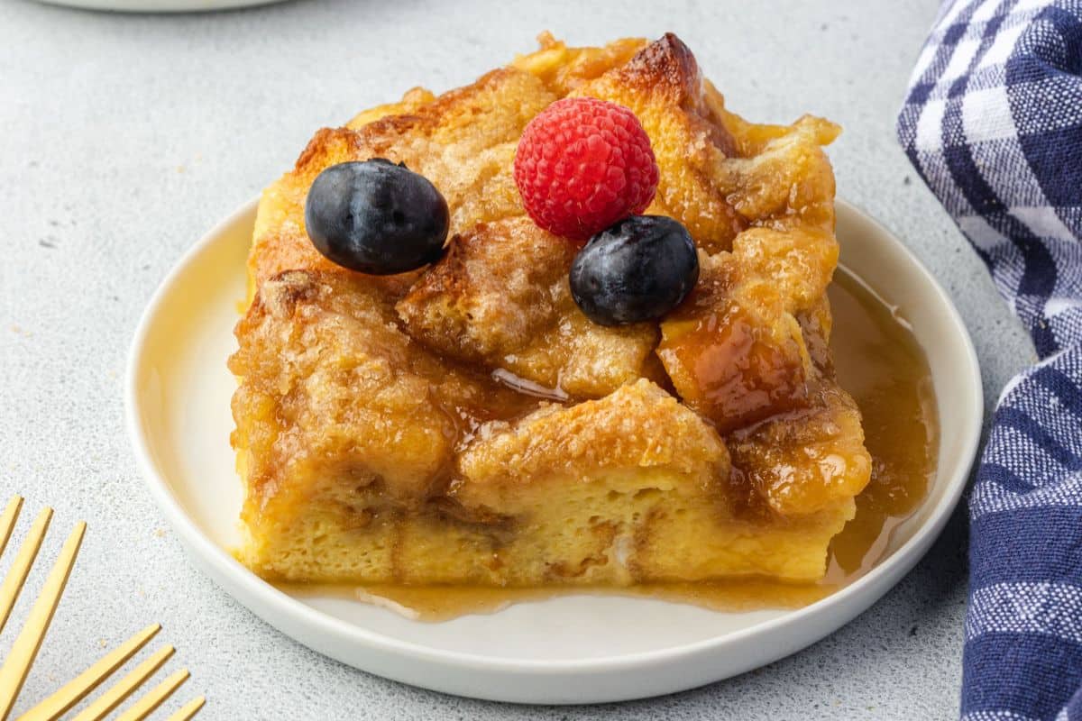 A slice of bread pudding topped with a raspberry, two grapes, and syrup on a white plate.