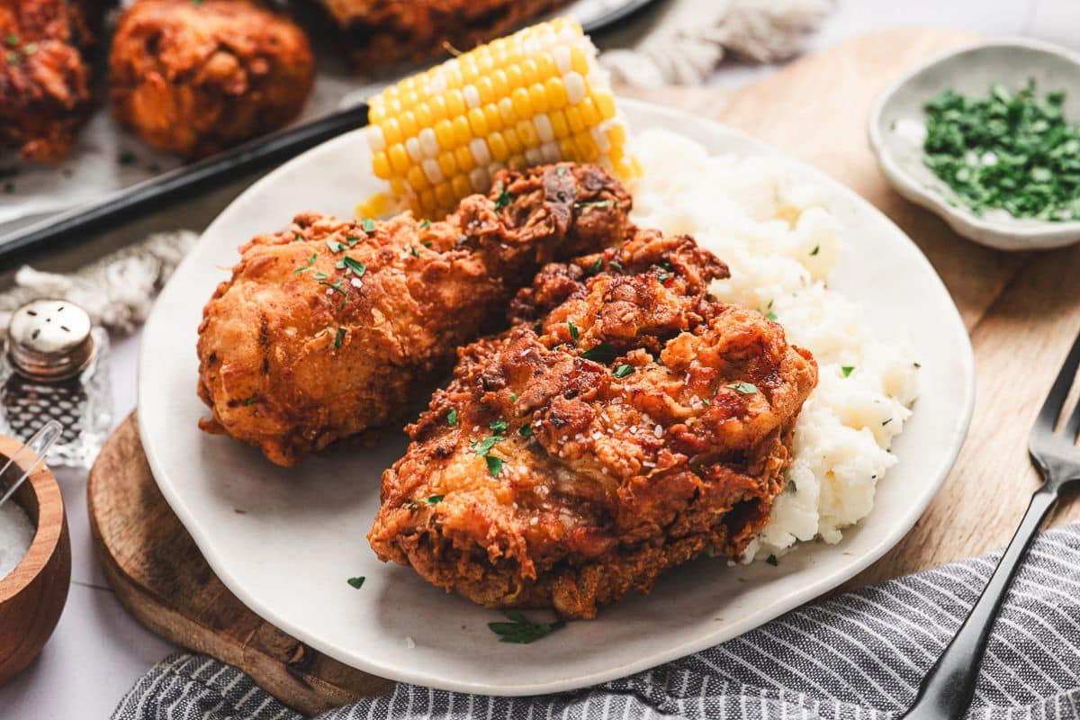 Plate with fried chicken, mashed potatoes, corn on the cob, and chopped herbs on a rustic table setting.