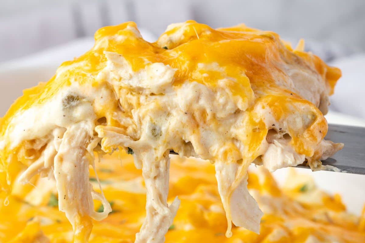 A spatula lifts a serving of cheesy chicken casserole with gooey melted cheddar cheese on top.
