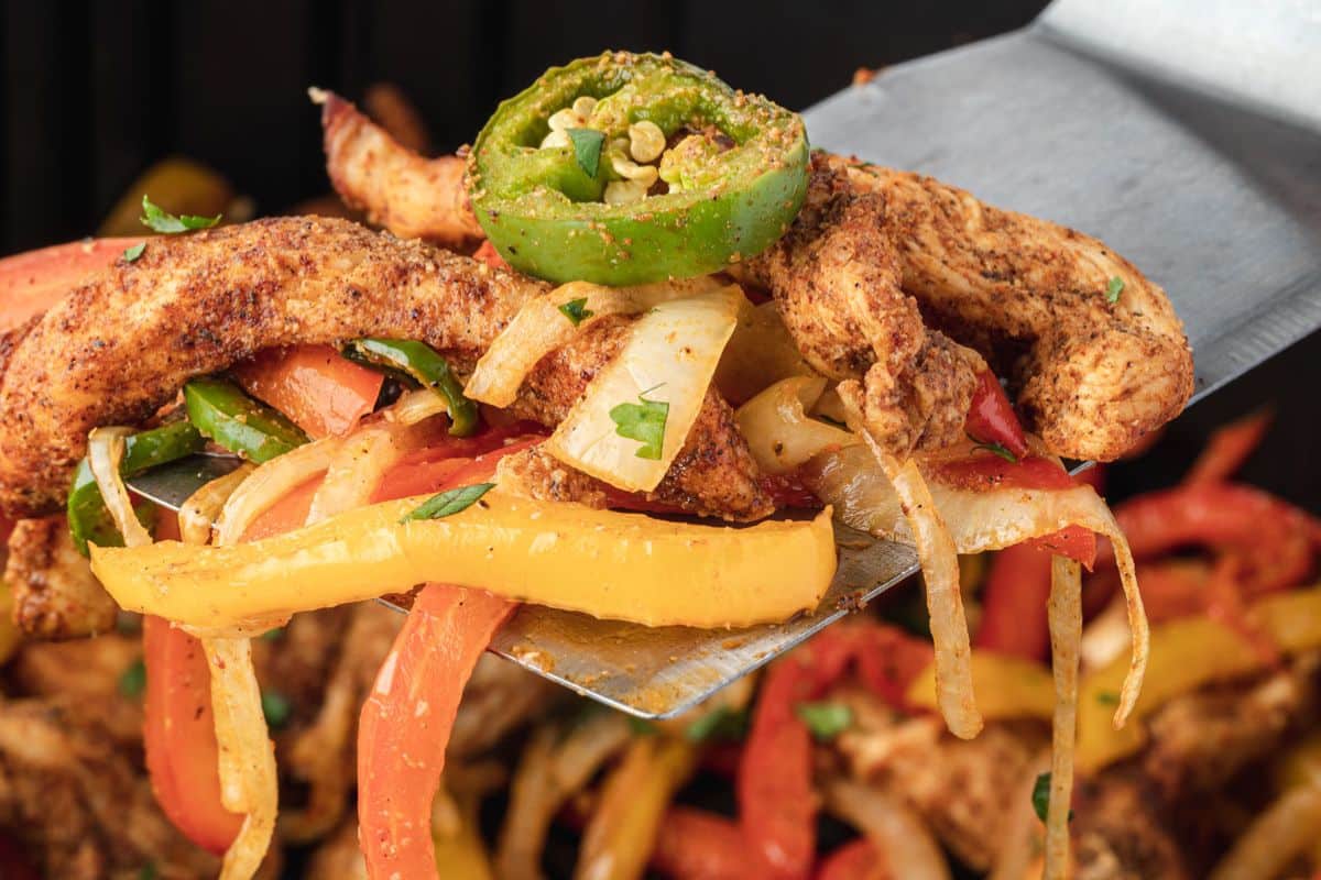Spatula holding seasoned chicken strips, bell peppers, onions, and sliced jalapeño, close up.