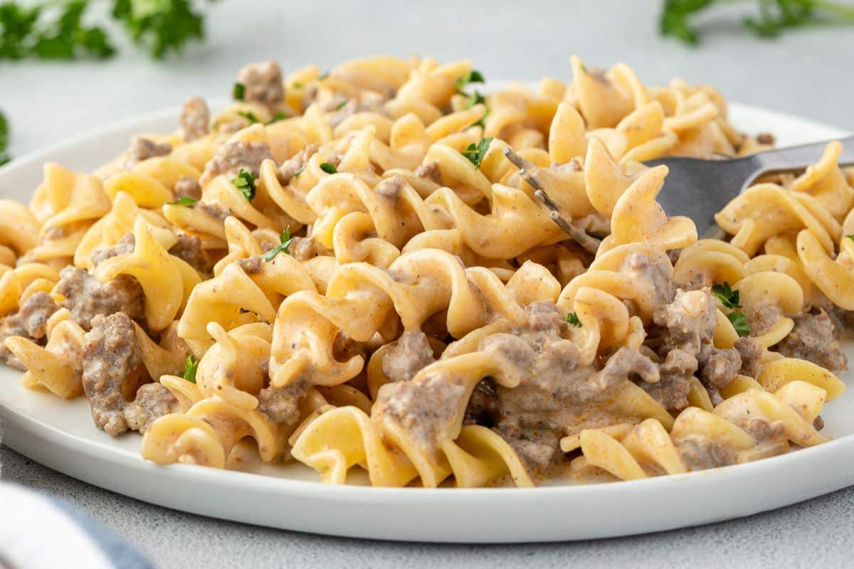 A plate of creamy beef stroganoff with rotini pasta, garnished with parsley, and a fork beside it.