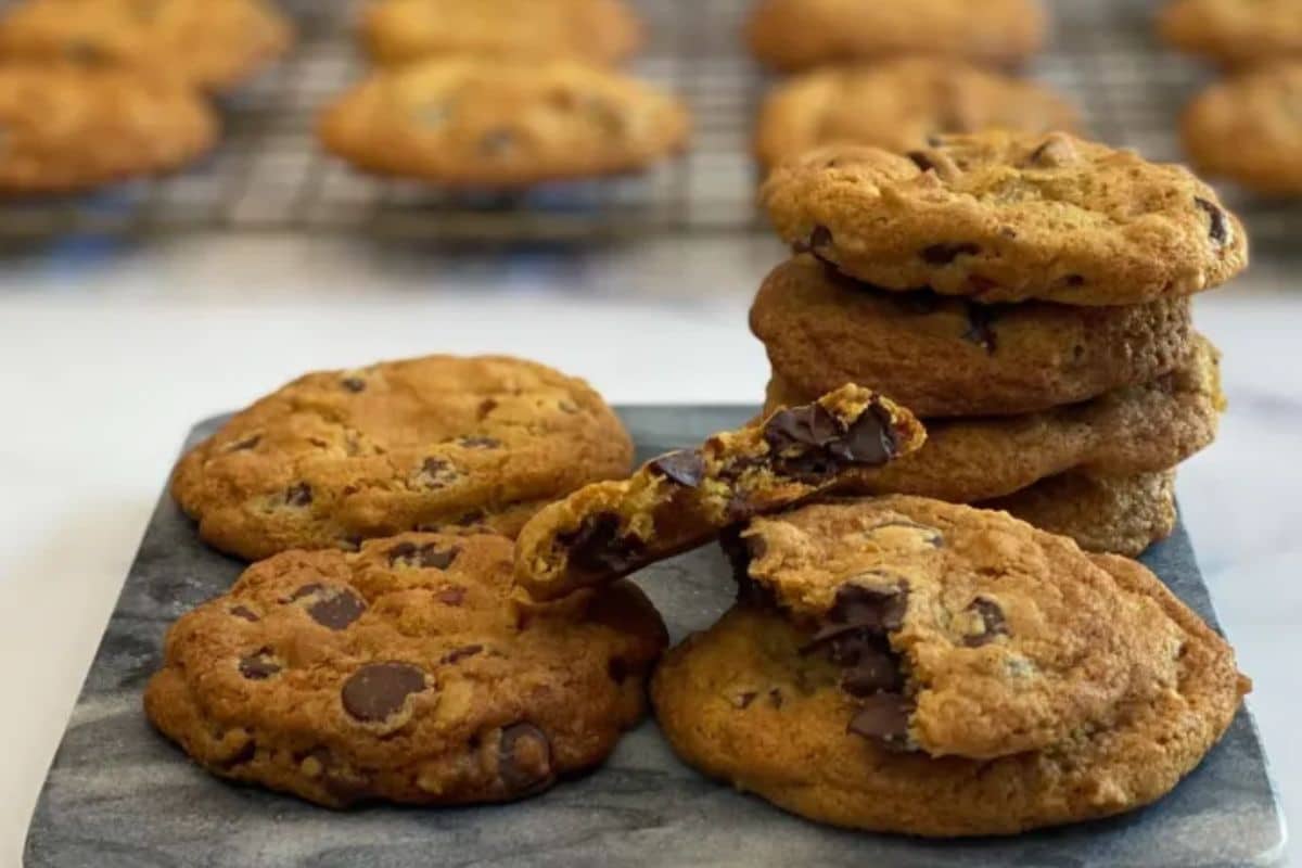 A stack of chocolate chip cookies with some broken pieces on a marble surface, more cookies in the background.