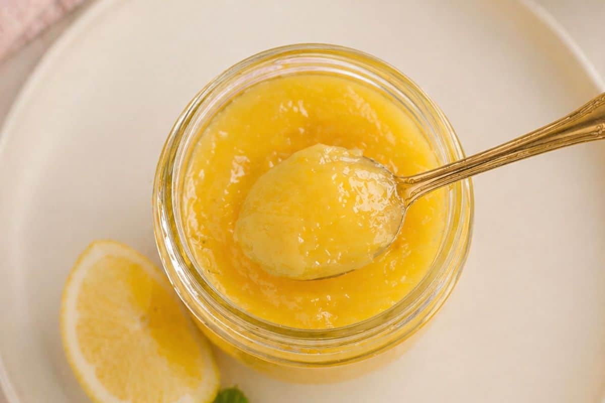 A spoon scoops lemon curd from a glass jar, with a lemon slice beside it on a white plate.