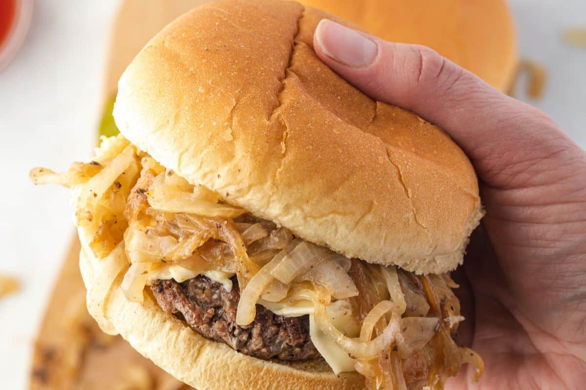 A hand holding a hamburger topped with sautéed onions and melted cheese in a soft bun.