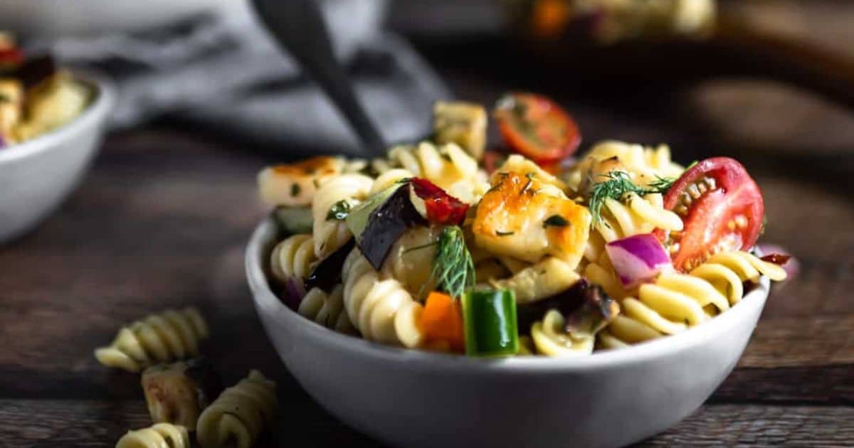 Bowl of pasta salad with rotini, cherry tomatoes, red onion, and herbs on a wooden table.