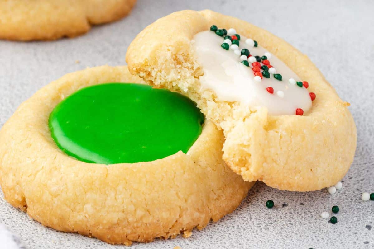 Two thumbprint cookies, one with green icing and one with white icing and sprinkles; one has a bite taken out.
