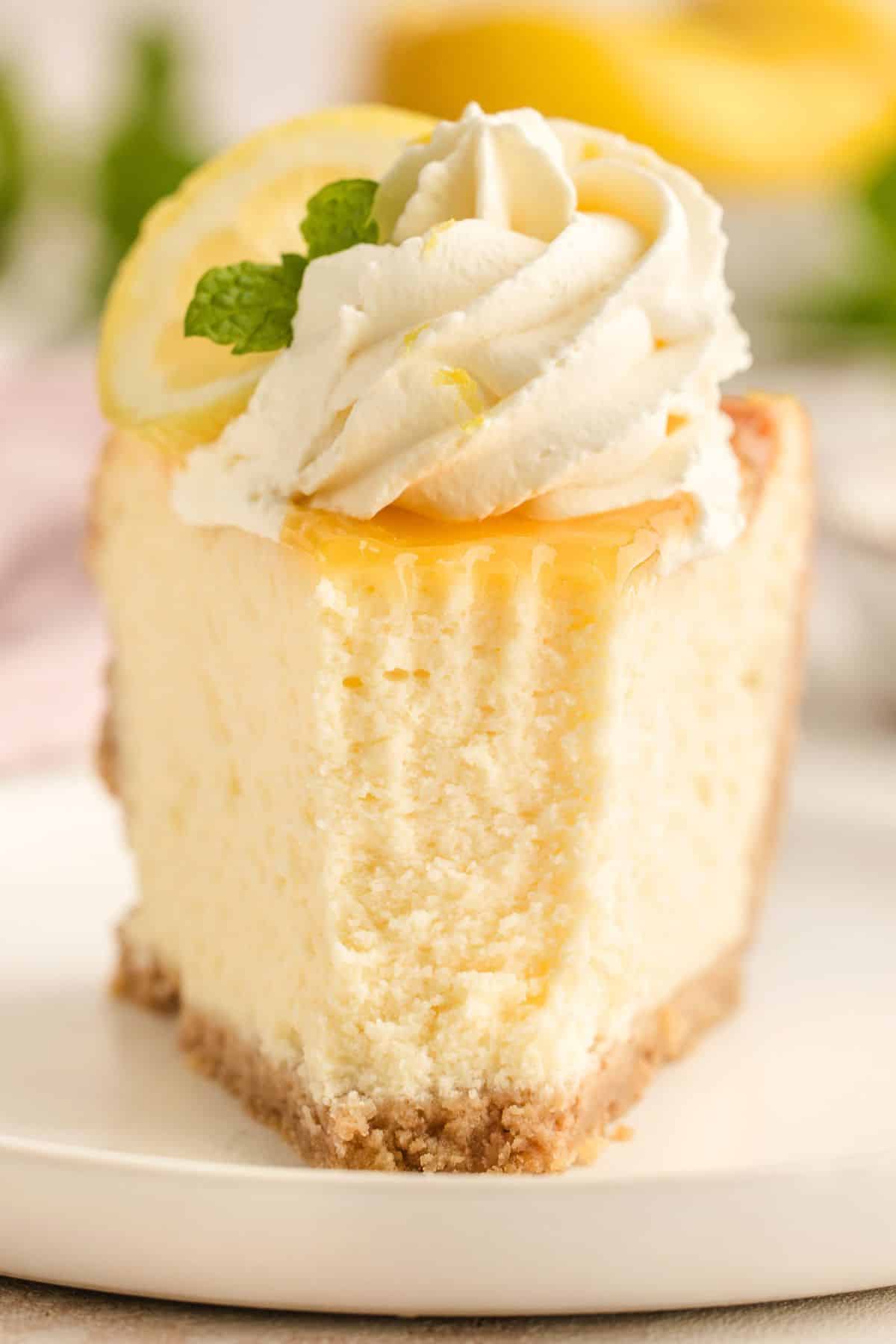 A slice of cheesecake topped with whipped cream, a lemon slice, and a mint leaf on a white plate.