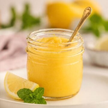 A jar of lemon curd spread with a spoon, garnished with a lemon slice and mint on a white plate.