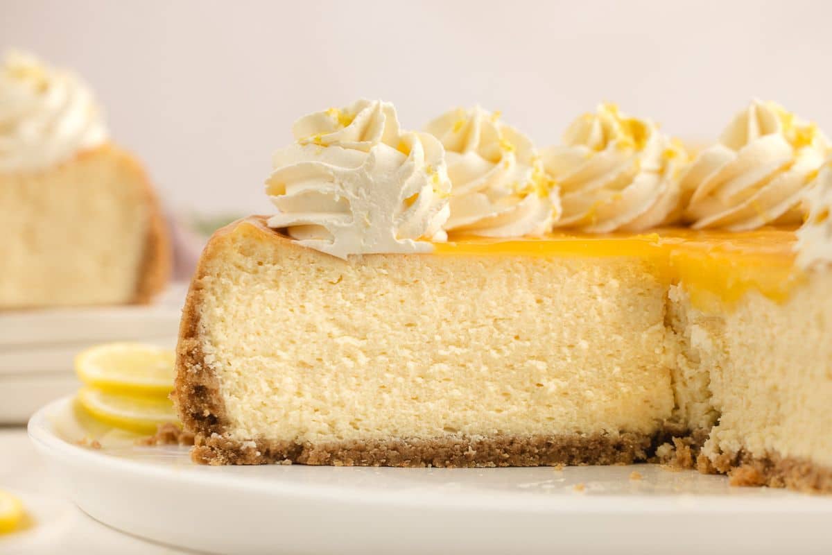 A slice of creamy cheesecake topped with whipped cream and lemon garnish on a white plate.