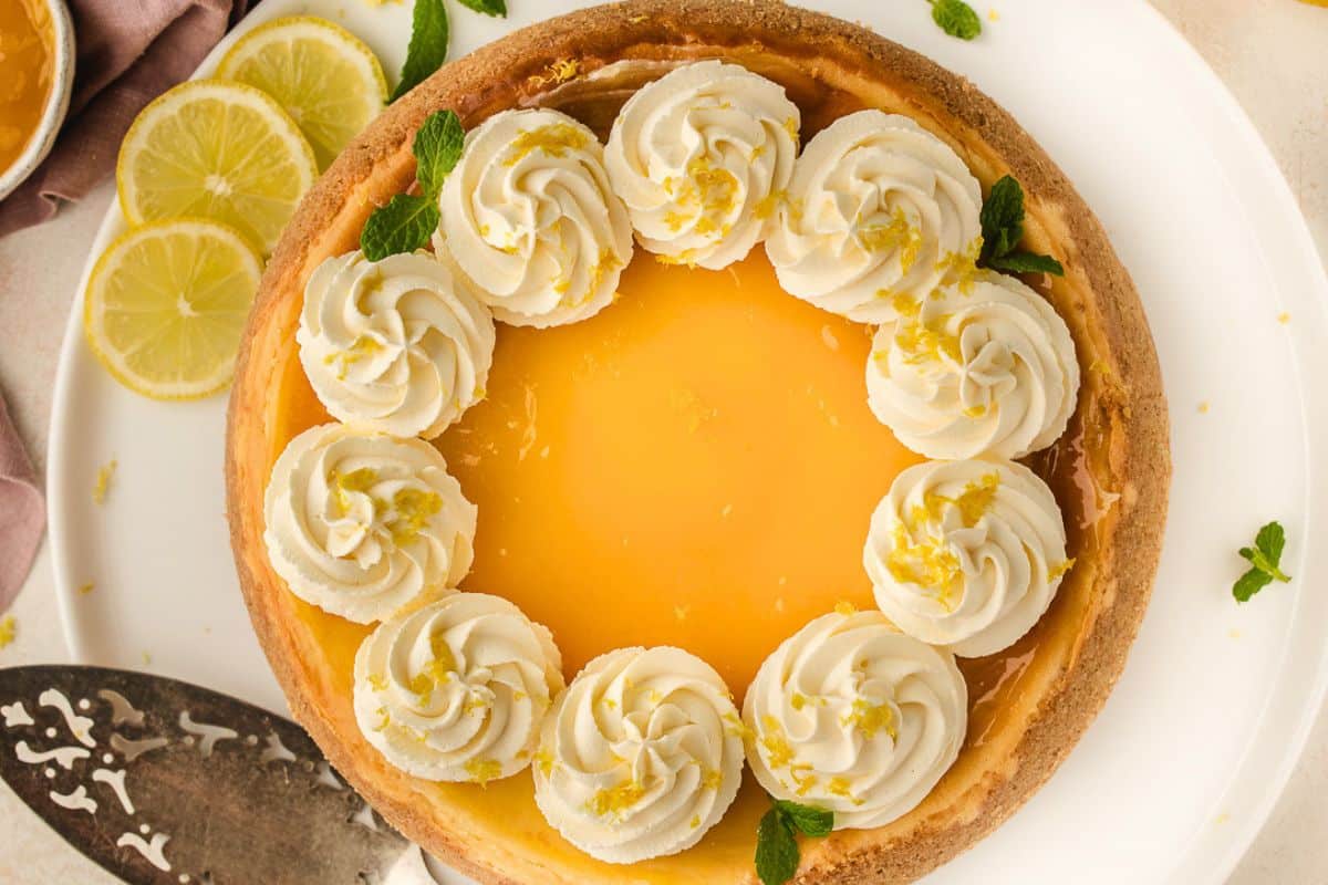 Lemon cheesecake topped with whipped cream swirls, lemon zest, and mint leaves, with lemon slices on the side.