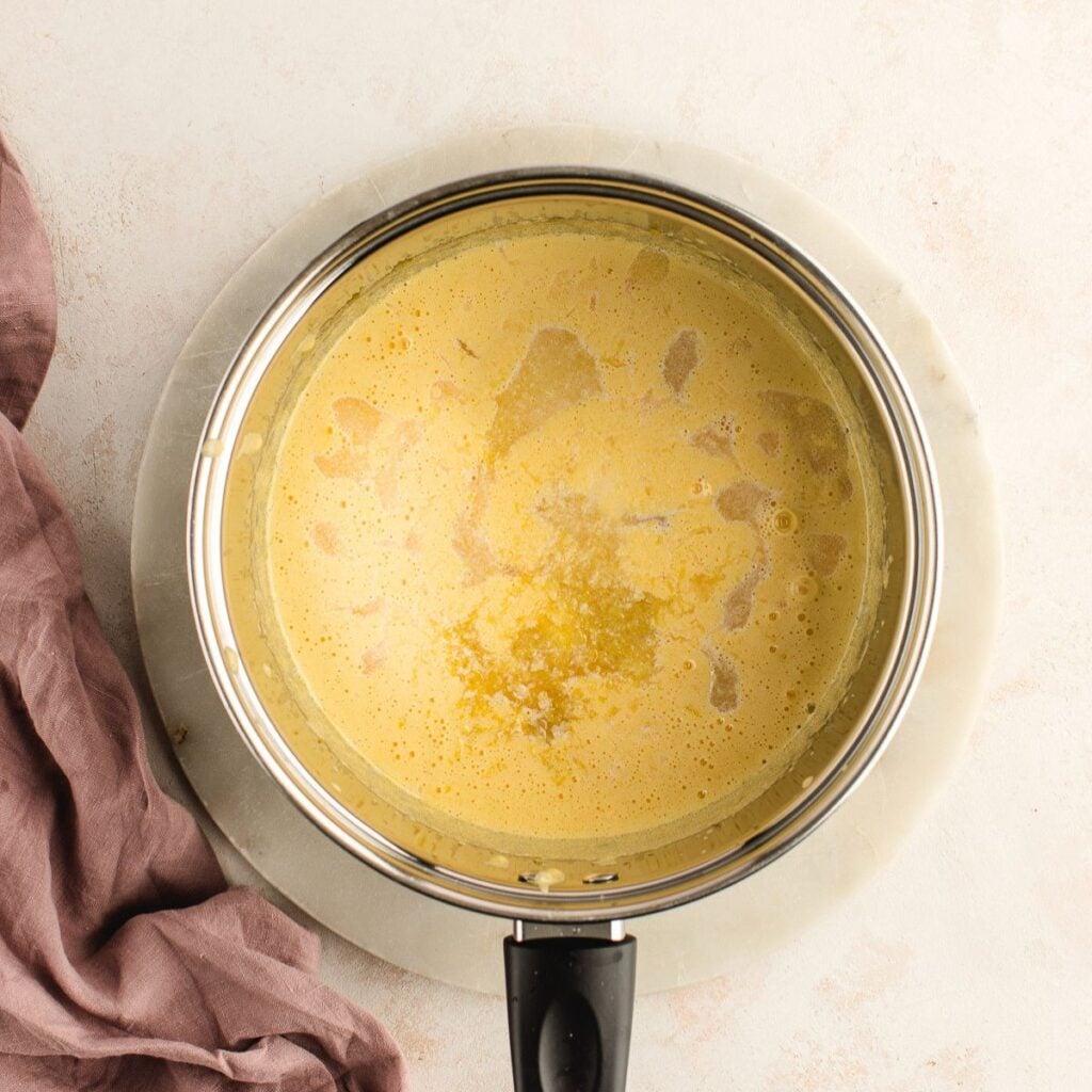 A saucepan filled with creamy yellow sauce sits on a round marble board next to a mauve cloth.