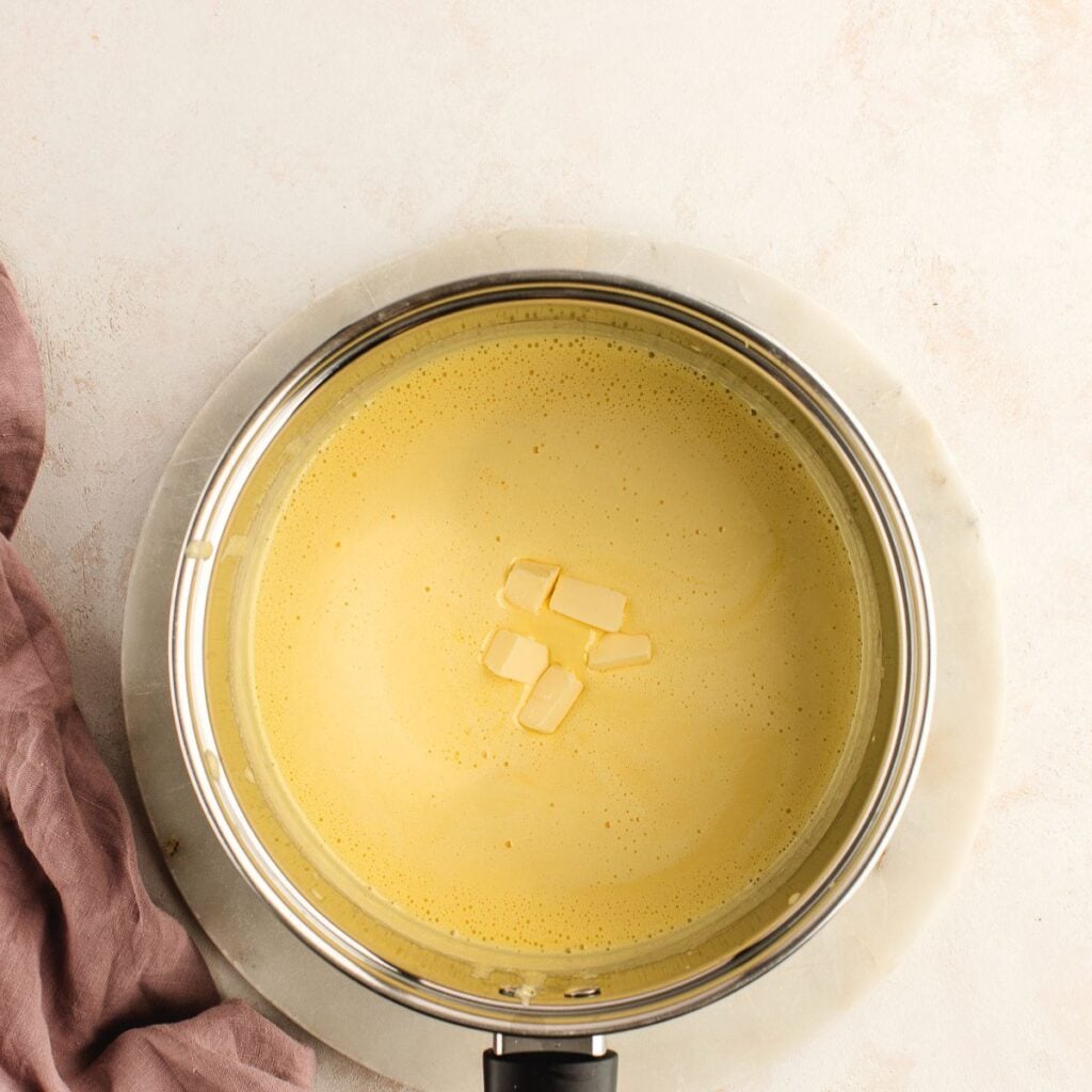 A pot of creamy yellow soup with small pieces of butter melting on top, placed on a light surface.