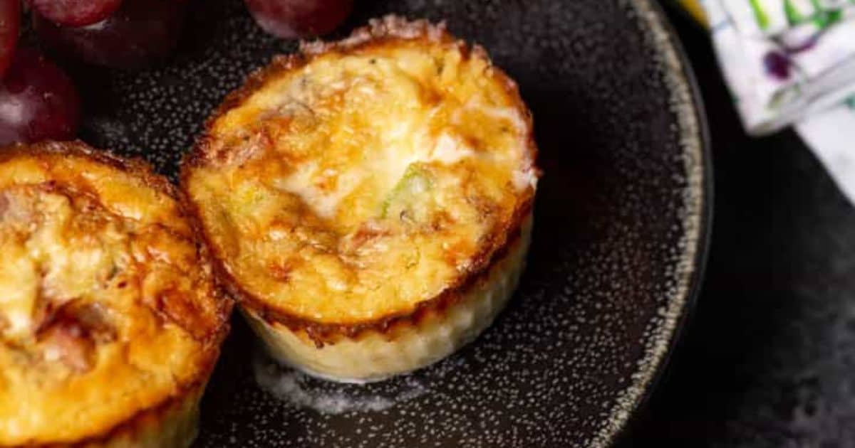 Two mini quiches on a dark plate, served with red grapes, viewed from above.