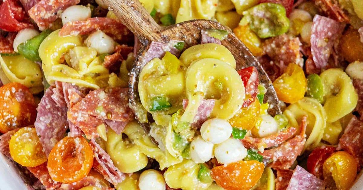 Wooden spoon scooping Italian tortellini pasta salad with salami, tomatoes, mozzarella, and peppers.