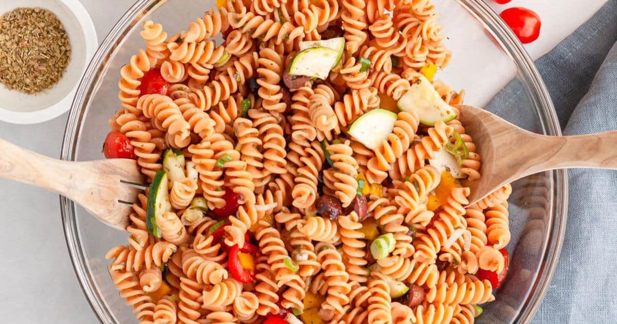 A bowl of rotini pasta salad with zucchini, tomatoes, olives, and wooden serving spoons.