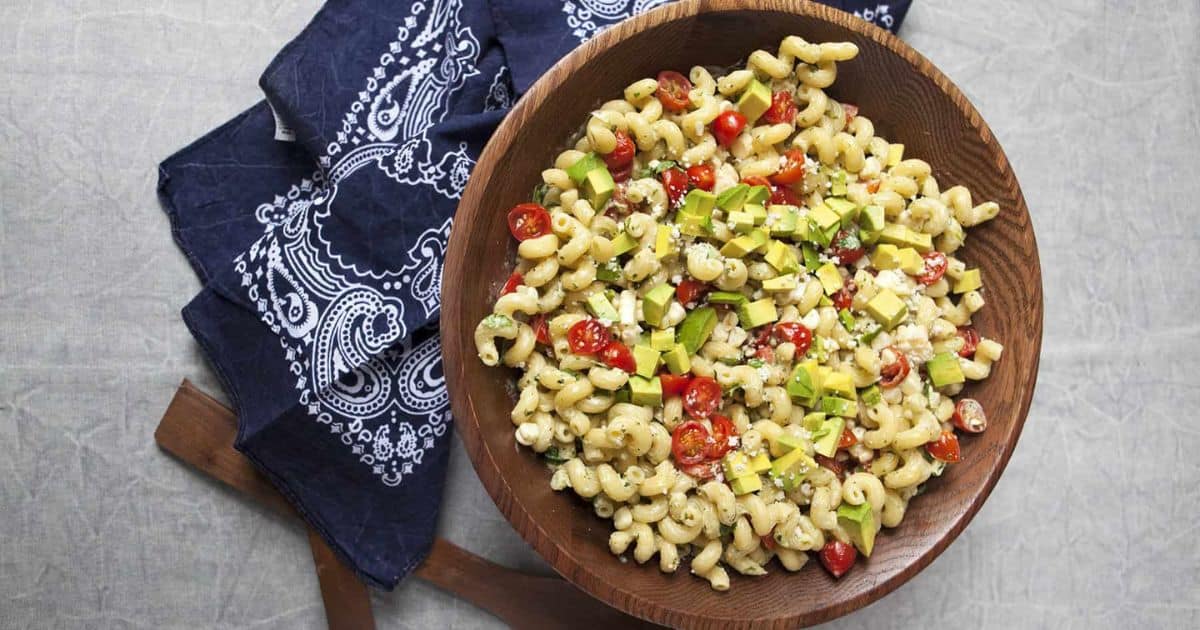 A wooden bowl of pasta salad with tomatoes, avocado, and cheese on a blue patterned napkin.