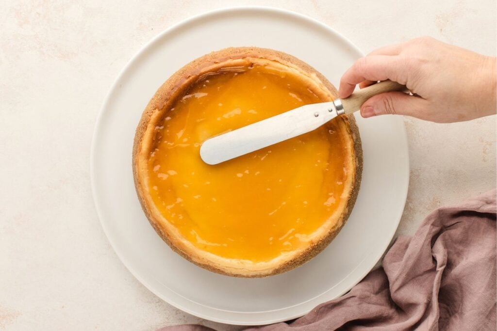 A hand spreads yellow glaze on a round cheesecake using a spatula on a white plate.