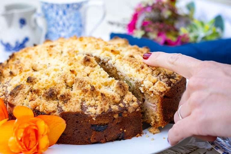 A hand with red nail polish points to a sliced crumb cake on a white plate, with flowers nearby.