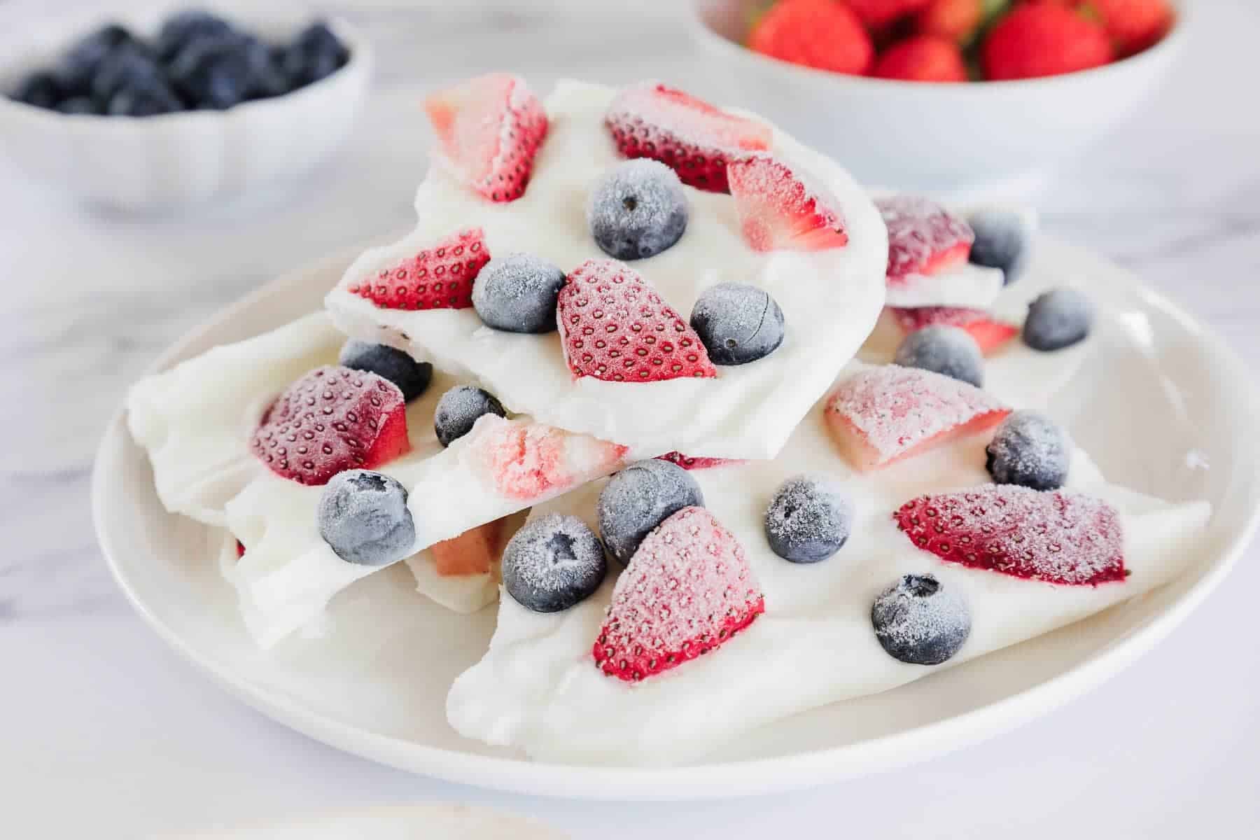 White yogurt bark topped with sliced strawberries and blueberries on a white plate, with berries in bowls behind.