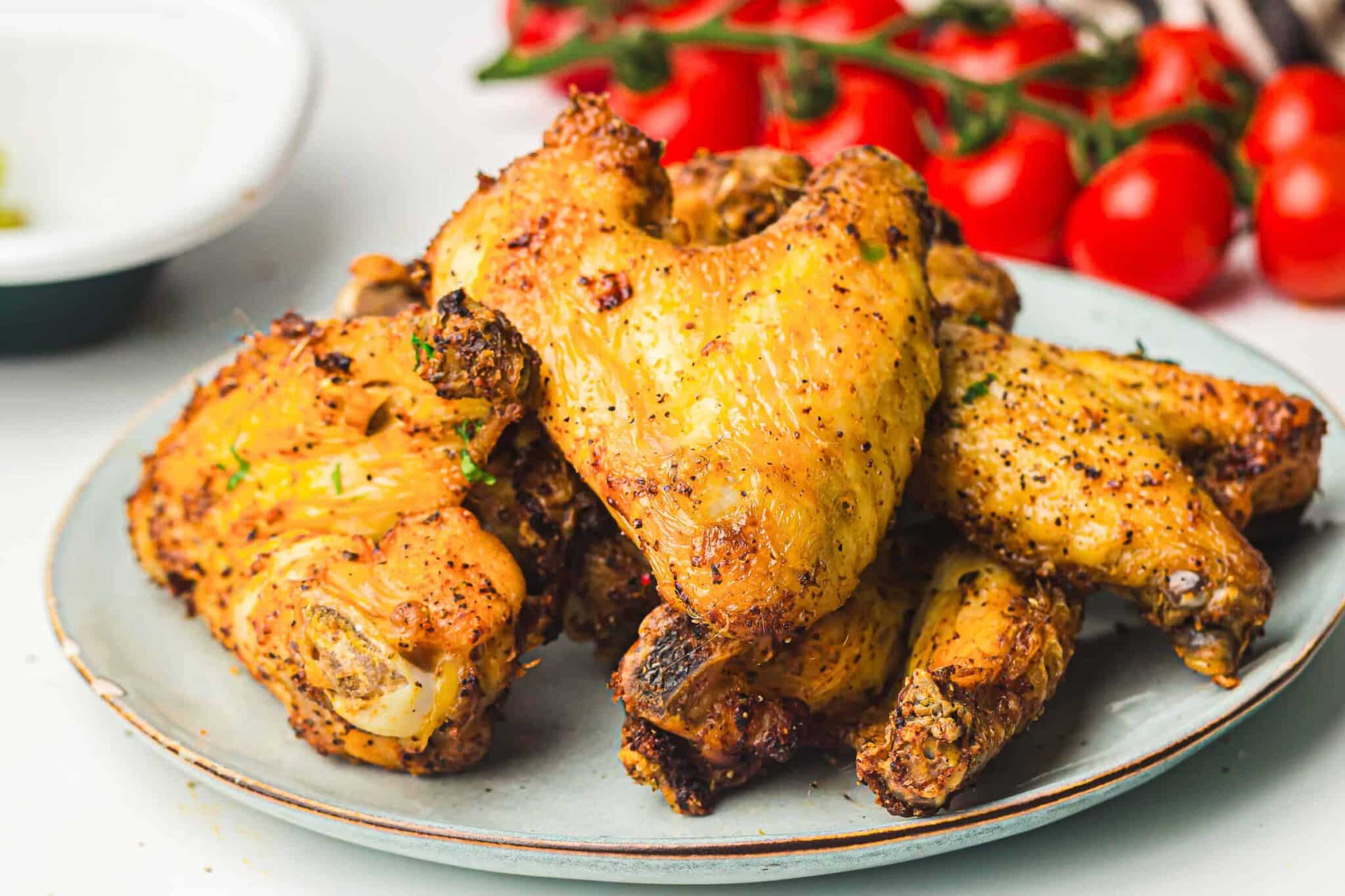 A plate of seasoned, cooked chicken wings with tomatoes on the vine in the background.