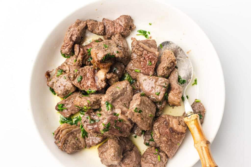 Cooked steak bites garnished with chopped herbs in a white bowl with a spoon.