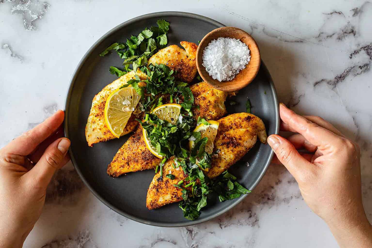 Grilled chicken breasts with lemon slices and herbs on a black plate, served with a bowl of coarse salt.