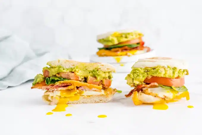 Breakfast sandwiches with bacon, egg, tomato, cheese, and avocado on English muffins, with yolk oozing out.