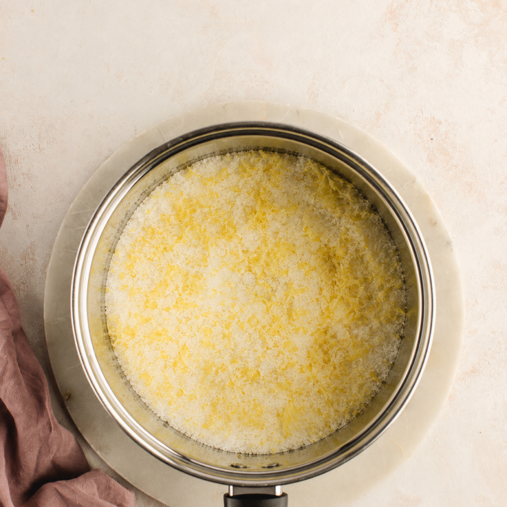 A metal pot filled with sugar and lemon zest sitting on a marble surface with a mauve cloth nearby.