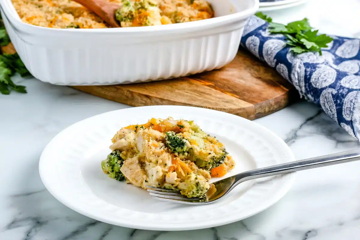 A serving of broccoli and chicken casserole on a white plate with a fork, casserole dish in the background.