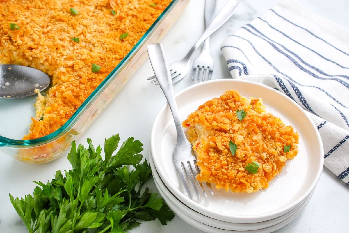 A serving of cheesy casserole on a white plate with parsley, stacked plates, and a baking dish nearby.