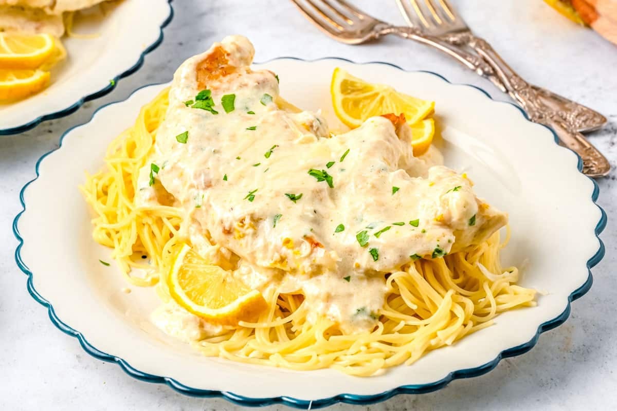 Chicken in creamy sauce over spaghetti, garnished with parsley and lemon wedges on a white plate.