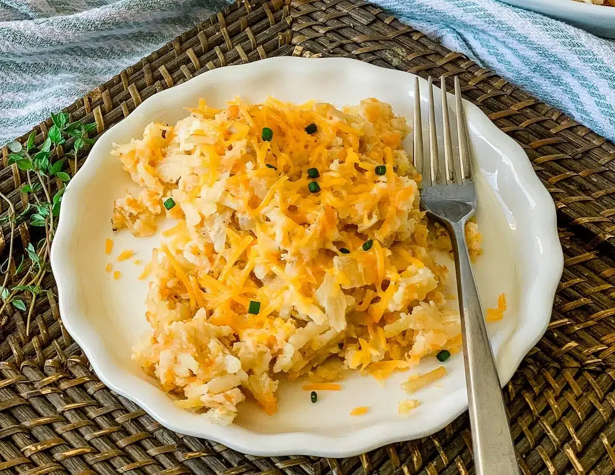 A plate of cheesy hashbrown casserole garnished with chopped chives, with a fork on a woven placemat.