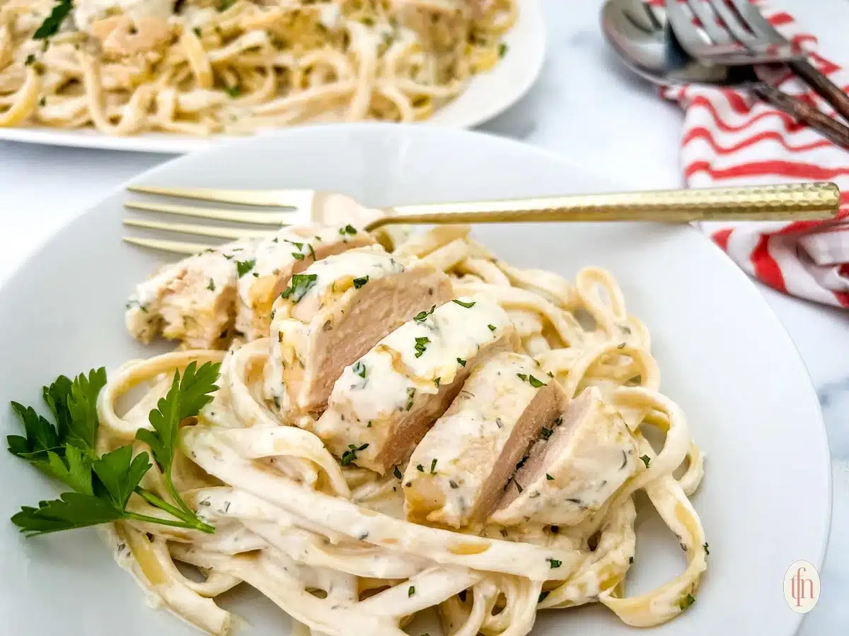 Plate of creamy fettuccine Alfredo with sliced chicken and parsley garnish, with a fork on the side.