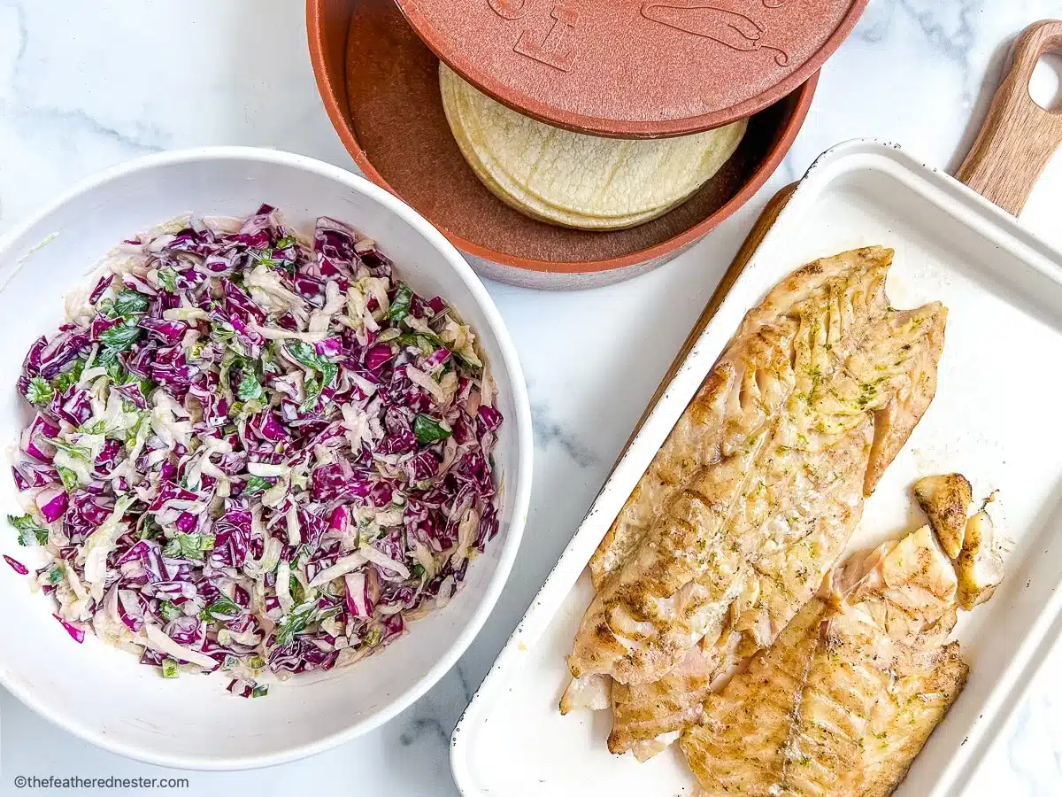 A bowl of cabbage slaw, cooked fish fillets, and a stack of corn tortillas on a white surface.