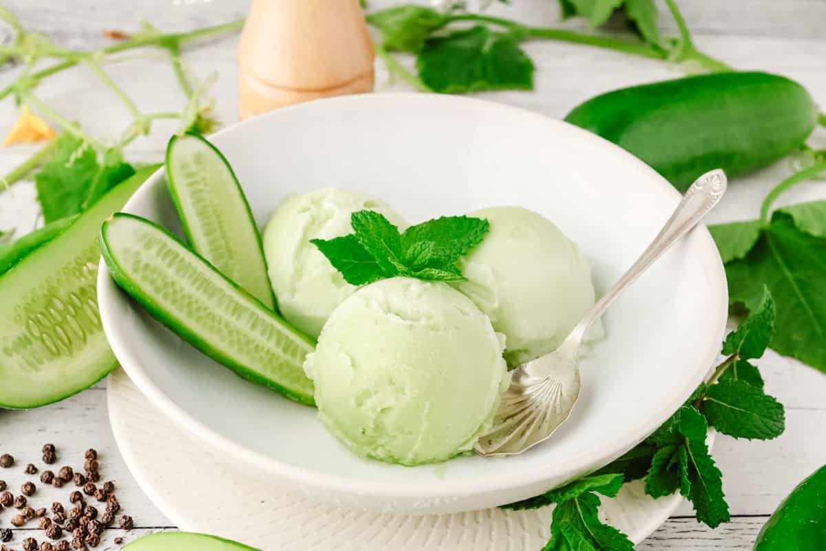 Three scoops of green ice cream in a bowl, garnished with cucumber slices and mint leaves, with a spoon.