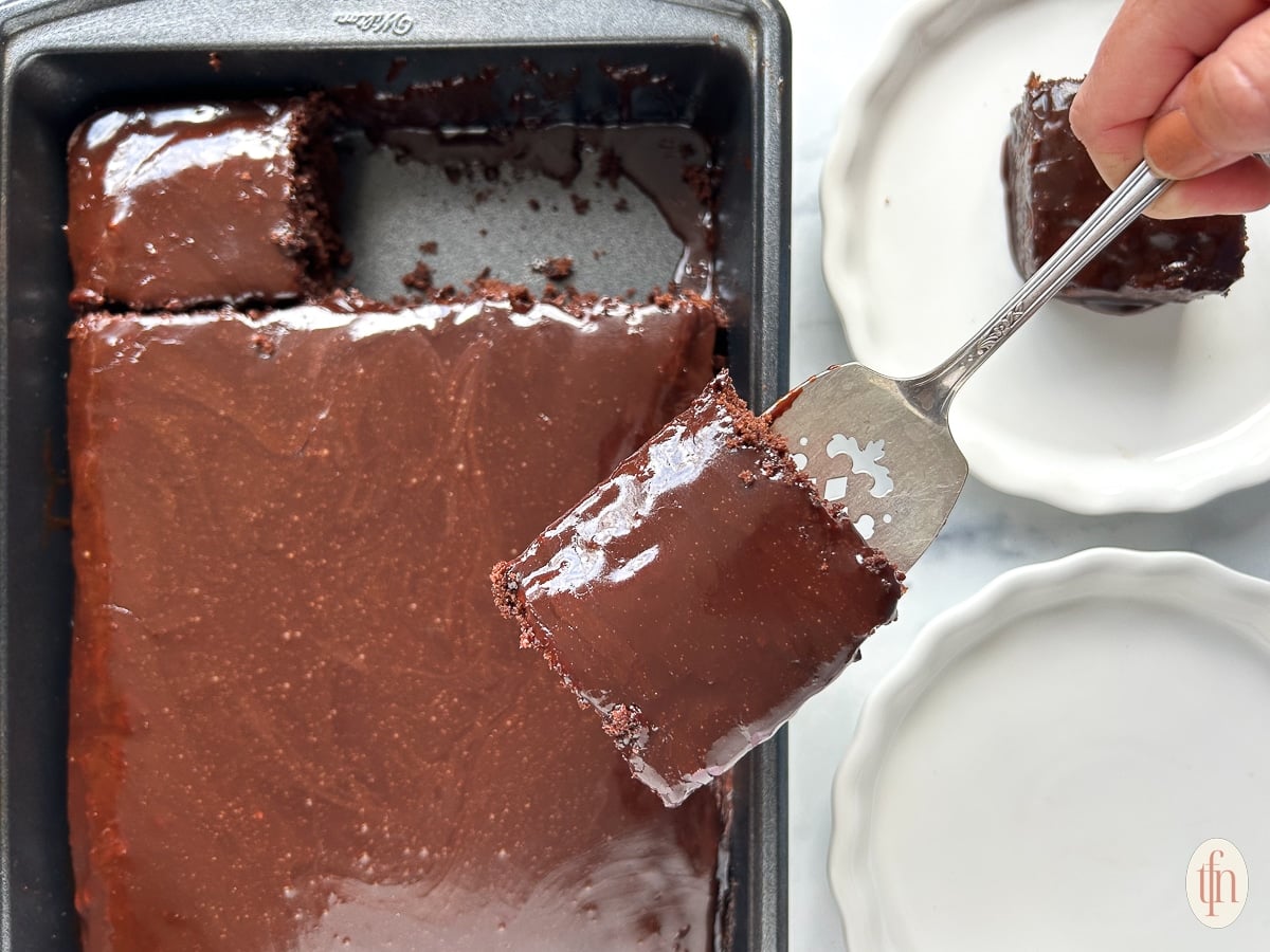 A hand lifts a slice of glossy chocolate cake from a pan, above two empty white dishes.