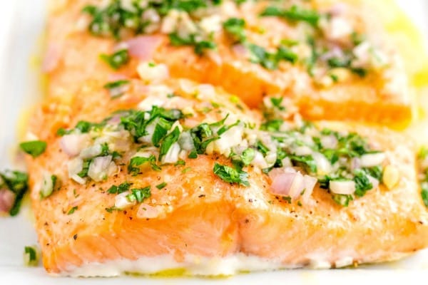 Two salmon fillets topped with chopped herbs, onions, and garlic, drizzled with olive oil on a white plate.