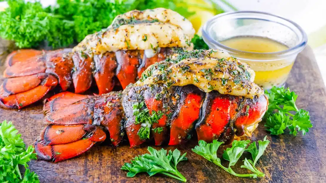 Two grilled lobster tails garnished with herbs, served with parsley and a small bowl of melted butter.