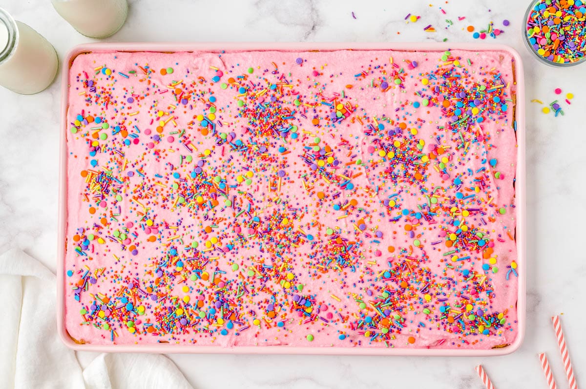A sheet cake with pink frosting and colorful sprinkles, next to two bottles of milk and striped straws.