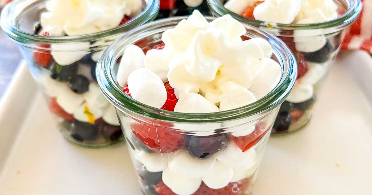 Three glass jars filled with blueberries, strawberries, mini marshmallows, and topped with whipped cream.