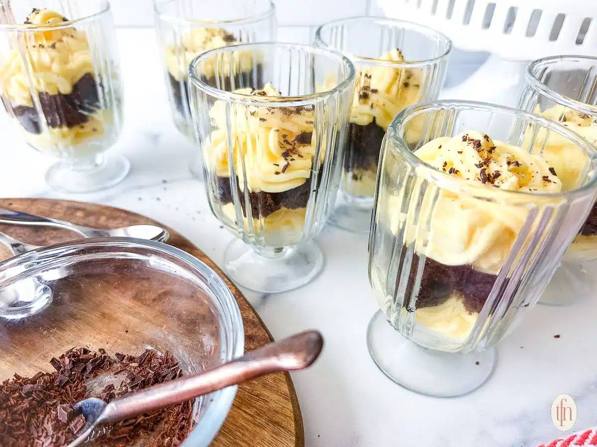 Six glass cups filled with layered chocolate cake and yellow custard, with grated chocolate on top.