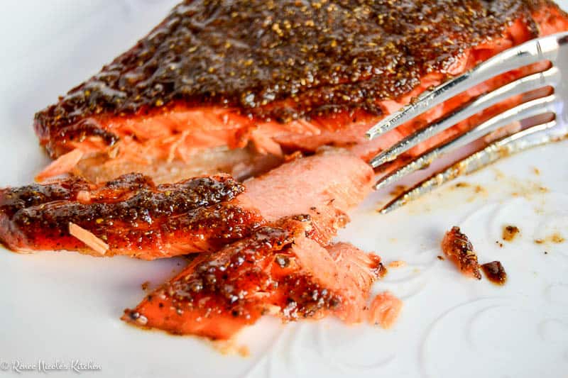 A fork flakes a piece of seasoned, cooked salmon on a white plate.