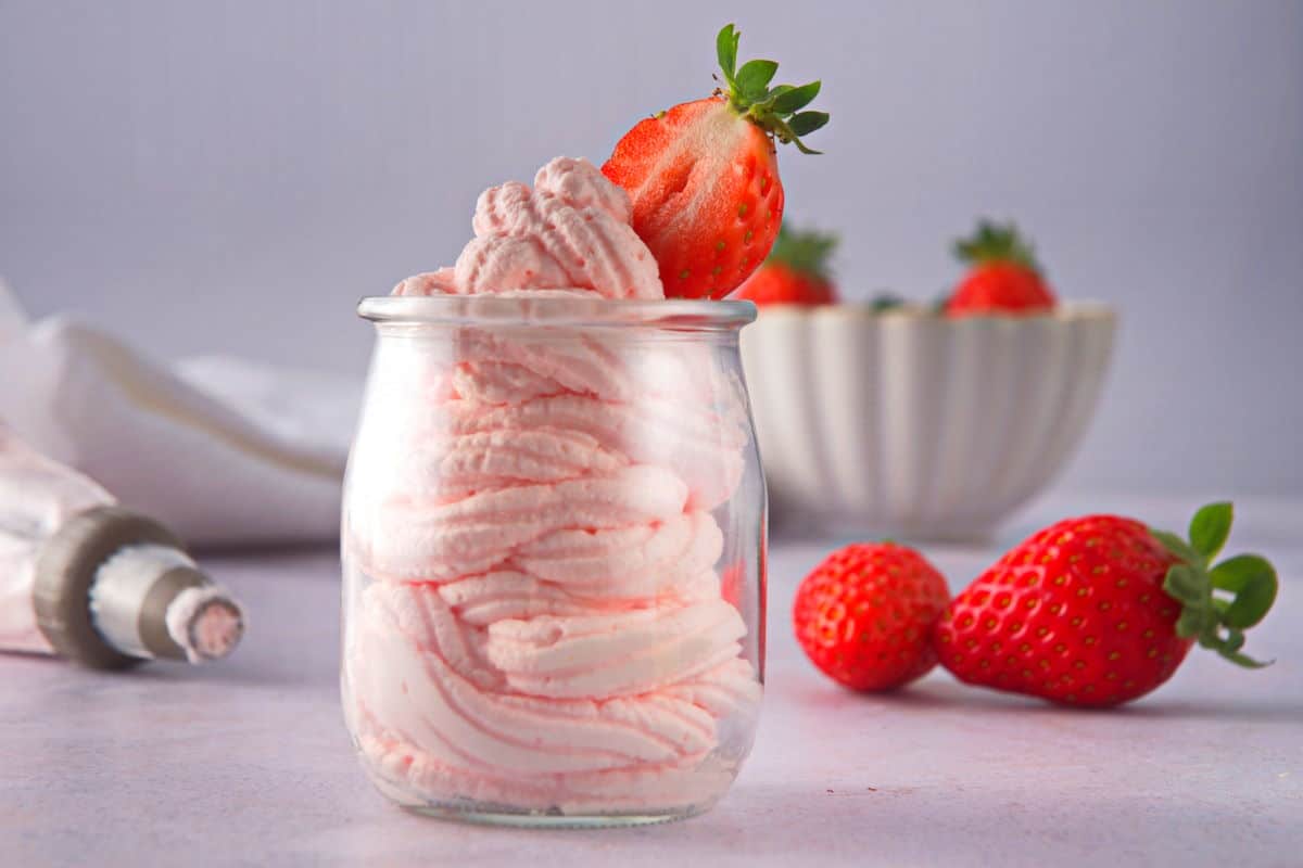 A glass jar filled with pink strawberry mousse, topped with a fresh strawberry, with more strawberries nearby.