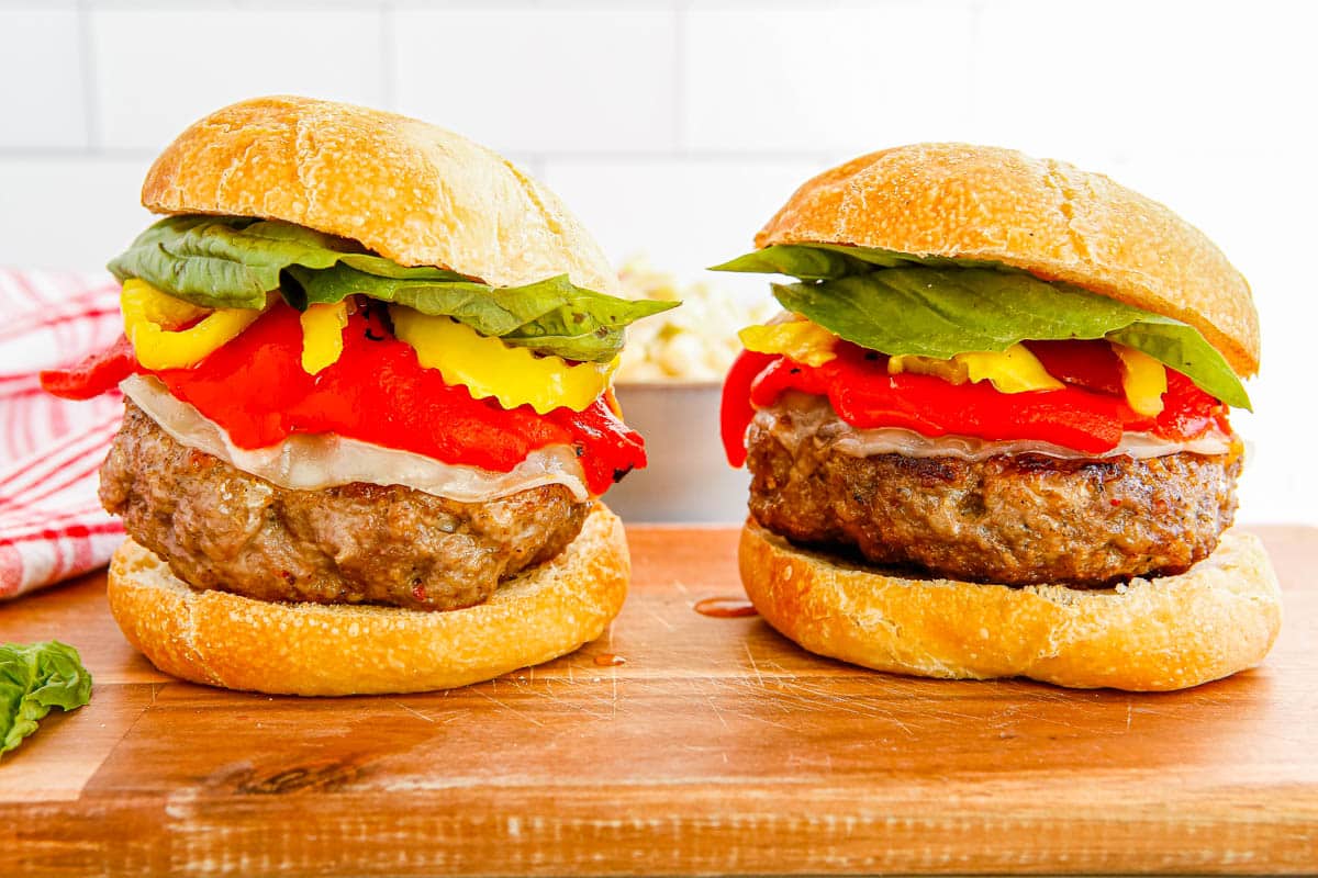 Two gourmet burgers with lettuce, cheese, roasted red pepper, and yellow pepper on a wooden board.