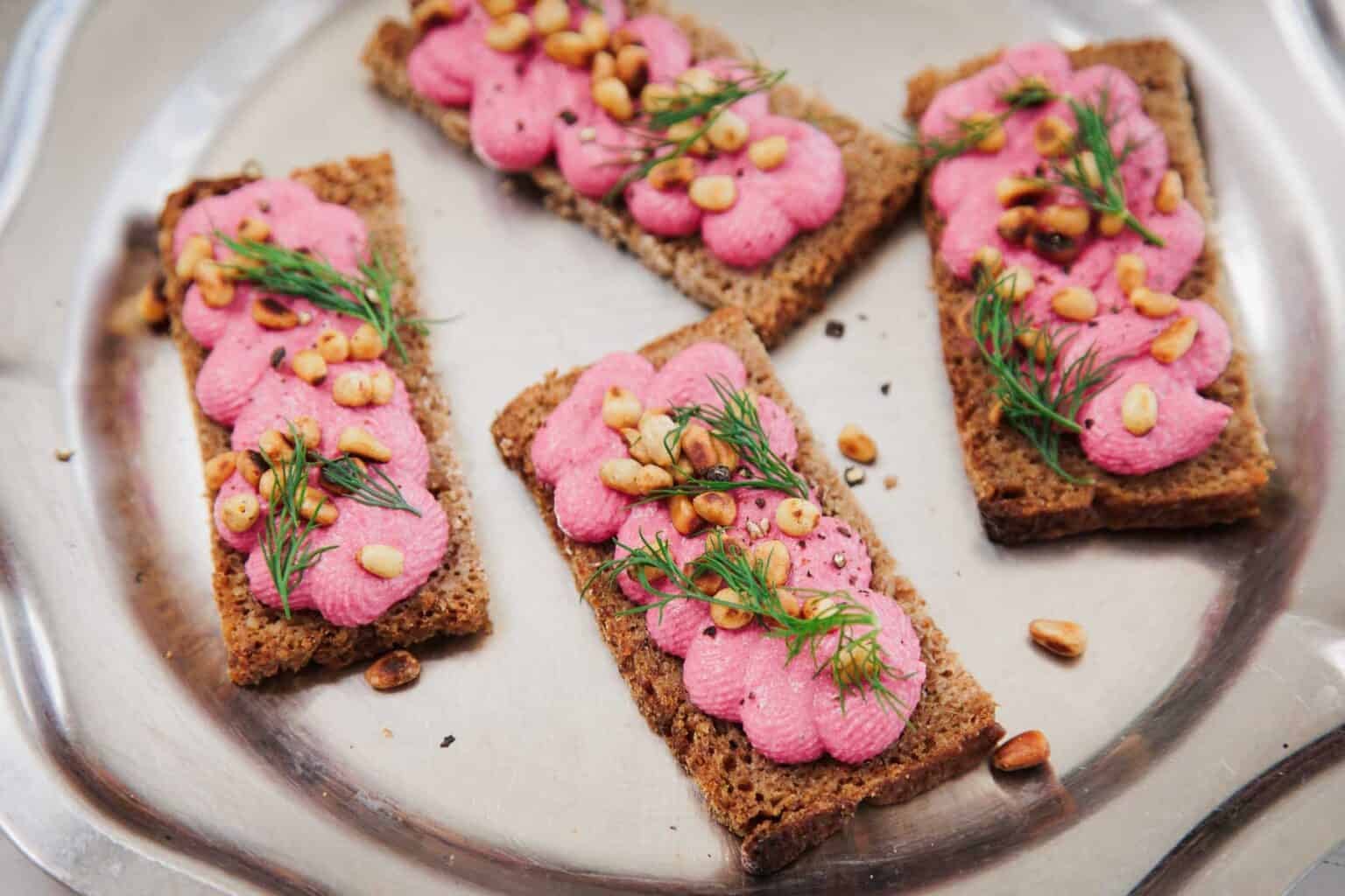 Four rye crispbreads topped with pink spread, pine nuts, and fresh dill on a silver tray.