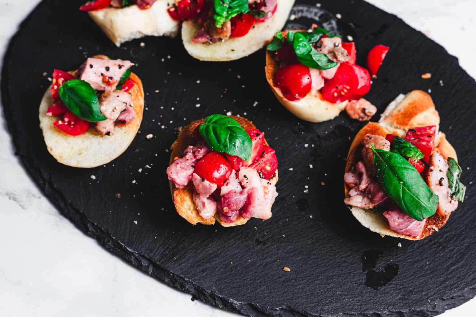 Slices of toasted bread topped with tomatoes, meat, and fresh basil on a black slate serving board.